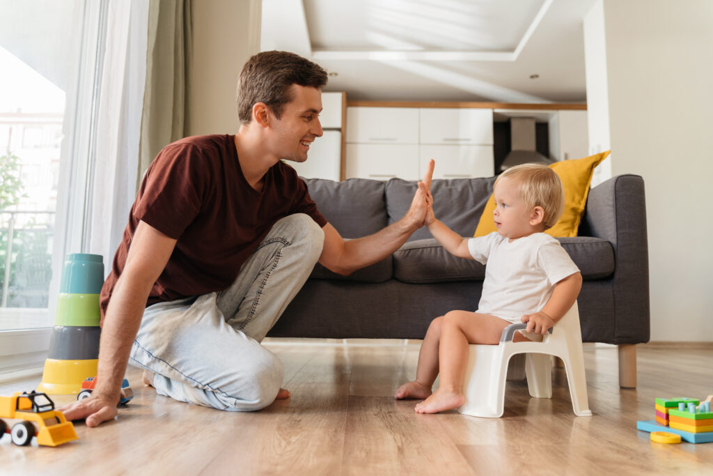 father assisting his little boy sitting on pot in potty training giving high five to his dad